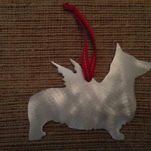 May include: A silver metal ornament shaped like a corgi with wings. The ornament has a red ribbon attached to the top.