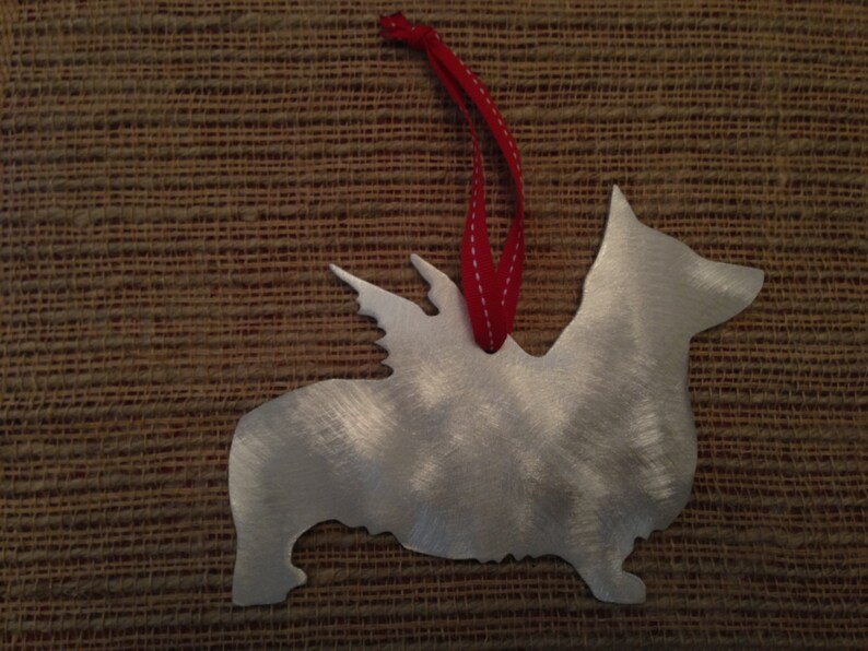 May include: A silver metal ornament shaped like a corgi with wings. The ornament has a red ribbon attached to the top.