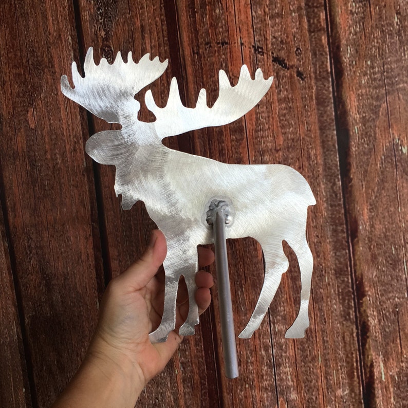 May include: A silver metal moose silhouette with a long metal rod attached to the back. The moose is standing on a wooden surface.