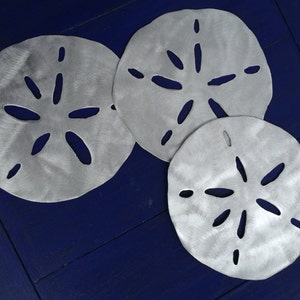 May include: Three silver metal sand dollar shapes on a dark blue wooden surface. The sand dollars are different sizes and have a cutout design.
