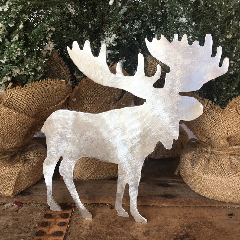 May include: A silver metal moose silhouette with a brushed finish. The moose is standing on a wooden surface with burlap fabric in the background.