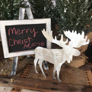 May include: A silver metal moose cutout with a chalkboard sign that reads "Merry Christ-Moose". The sign is on a white easel and the moose is on a wooden surface with a burlap sack behind it.