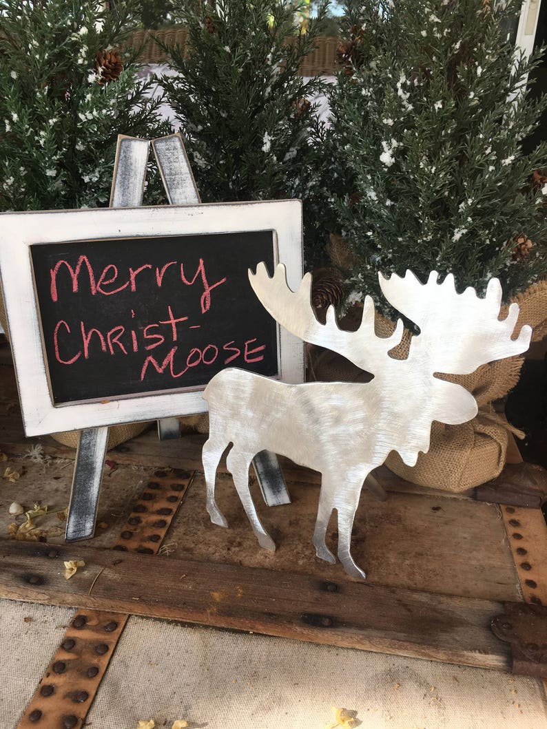 May include: A silver metal moose cutout with a chalkboard sign that reads "Merry Christ-Moose". The sign is on a white easel and the moose is on a wooden surface with a burlap sack behind it.
