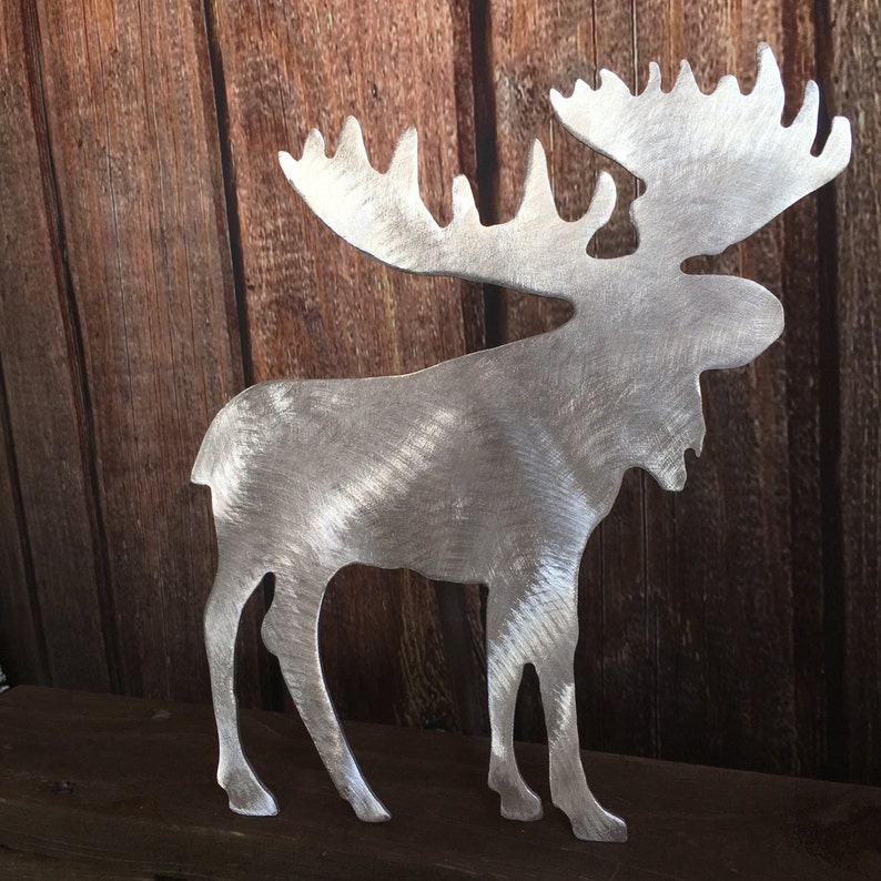 May include: A silver metal moose silhouette with a brushed finish. The moose is standing on a wooden surface.
