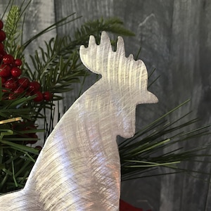 Puede incluir: Una silueta de gallo de metal plateado con acabado cepillado. El gallo est&aacute; mirando hacia la derecha y tiene una cresta y una papada detalladas.