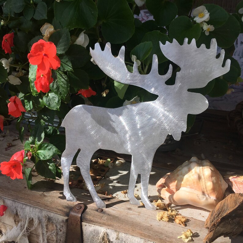 May include: A silver metal moose silhouette with a brushed finish. The moose is standing on a wooden surface with a background of green plants and red flowers.