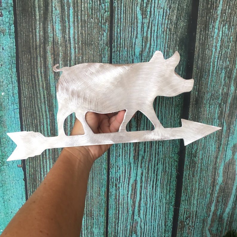 May include: A metal pig wind vane with an arrow pointing to the right. The pig is silver and the arrow is silver. The wind vane is held in a hand against a teal wooden background.
