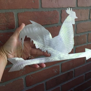 Puede incluir: Un veleta de metal plateado en forma de gallo con una flecha que apunta hacia la derecha. El gallo est&aacute; mirando hacia la izquierda y tiene un dise&ntilde;o detallado.