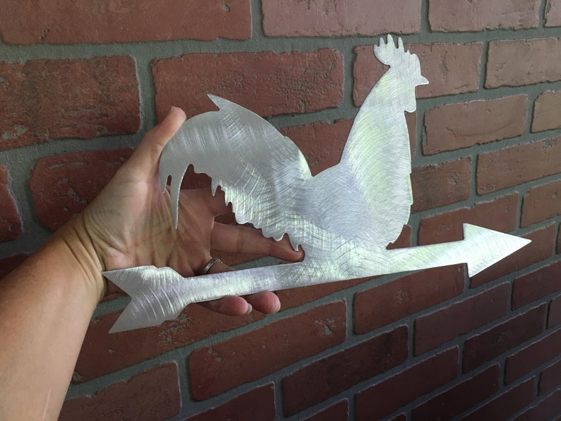 Puede incluir: Un veleta de metal plateado en forma de gallo con una flecha que apunta hacia la derecha. El gallo est&aacute; mirando hacia la izquierda y tiene un dise&ntilde;o detallado.