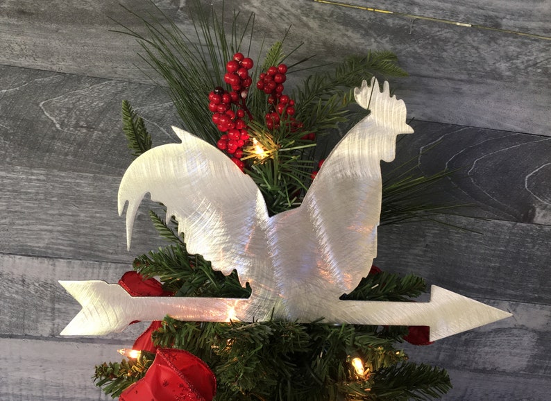 Puede incluir: Un adorno para &aacute;rbol de Navidad de metal plateado en forma de gallo con una flecha apuntando hacia la derecha. El gallo est&aacute; de pie en una rama de un &aacute;rbol de Navidad.