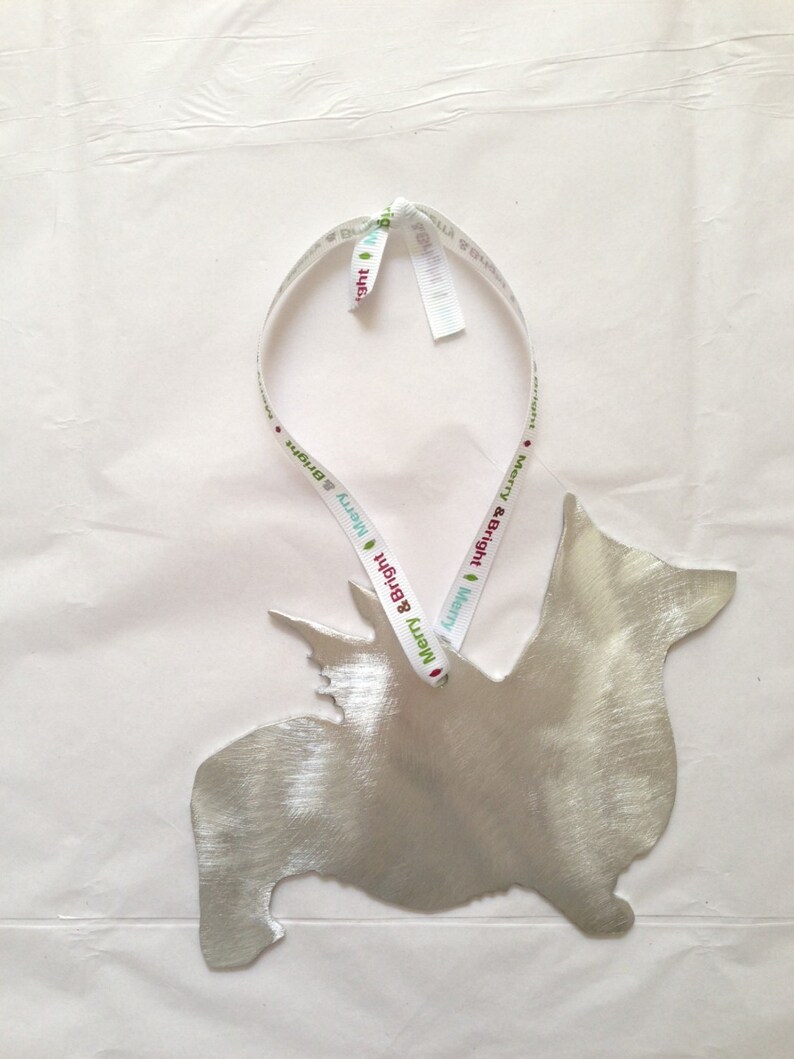 May include: A silver metal ornament shaped like a dog with a white ribbon tied around it. The ribbon has the text "Merry & Bright" printed on it.