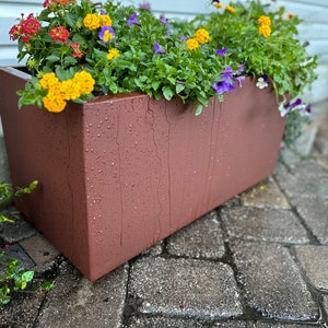 May include: A terracotta-colored planter with a variety of colorful flowers, including yellow, orange, red, and purple, planted in it. The planter is sitting on a gray brick patio.