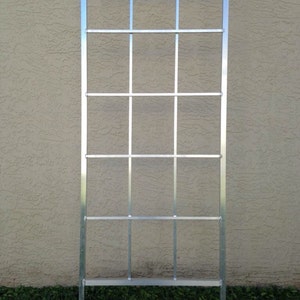May include: A silver metal trellis with a rectangular shape and a grid pattern. The trellis is standing upright on a grassy surface.