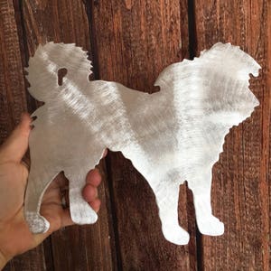 May include: A silver metal silhouette of a dog, possibly a breed like a Shih Tzu or a Bichon Frise, with a fluffy tail. The silhouette is cut out of metal and has a brushed finish.