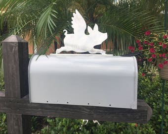 May include: A white metal mailbox with a pig with wings sculpture on top. The mailbox is mounted on a wooden post.