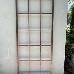 May include: A copper-coloured metal trellis with a rectangular frame and a grid pattern. The trellis is standing upright against a white wall.