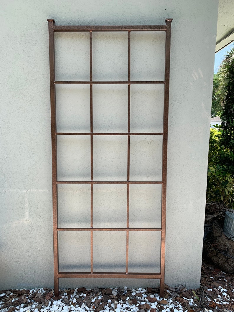 May include: A copper-coloured metal trellis with a rectangular frame and a grid pattern. The trellis is standing upright against a white wall.