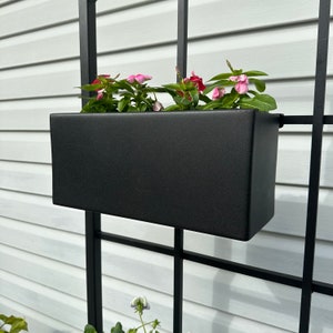 May include: A black metal planter box with pink flowers hanging on a black metal trellis against a white siding background.
