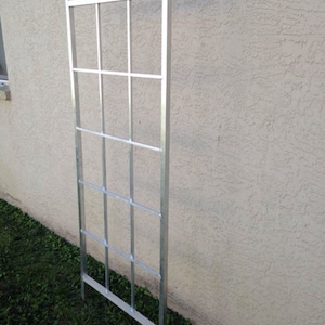 May include: A silver metal trellis with a rectangular frame and a grid pattern. The trellis is standing upright against a white wall.