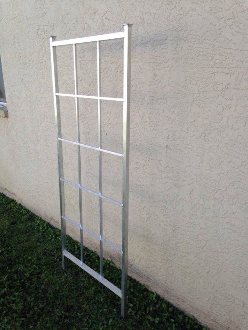 May include: A silver metal trellis with a rectangular frame and a grid pattern. The trellis is standing upright against a white wall.