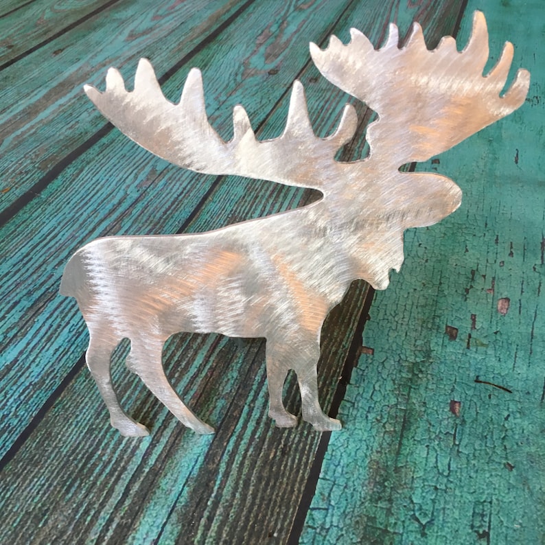 May include: A silver metal moose silhouette with a brushed finish. The moose is standing on a teal blue wooden surface.