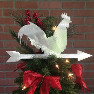 Puede incluir: Un adorno para &aacute;rbol de Navidad de metal blanco en forma de gallo con un lazo rojo y una flecha plateada. El gallo est&aacute; posado en la cima de un &aacute;rbol de Navidad verde con bayas rojas.