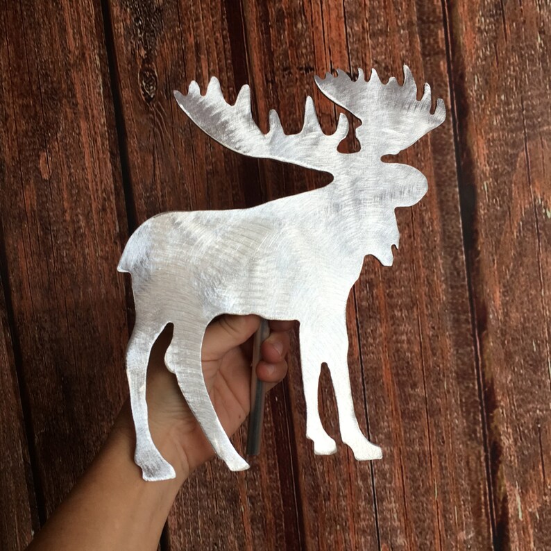 May include: A silver metal moose silhouette with a brushed finish. The moose is standing on a wooden surface.