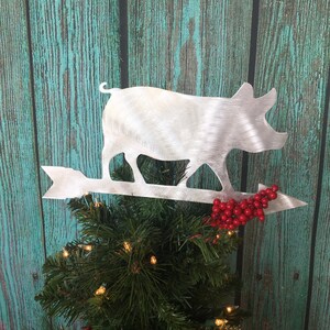 May include: A silver pig Christmas tree topper with a red berry sprig on an arrow. The pig is walking on the arrow and is facing the right side of the image.