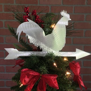 Puede incluir: Un adorno para &aacute;rbol de Navidad de metal plateado en forma de gallo con una flecha apuntando hacia la derecha. El gallo est&aacute; posado en la cima de un &aacute;rbol de Navidad verde con lazos rojos y luces blancas.