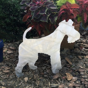 May include: A metal silhouette of a wirehaired fox terrier dog. The silhouette is a light silver color and is standing on a bed of brown mulch.