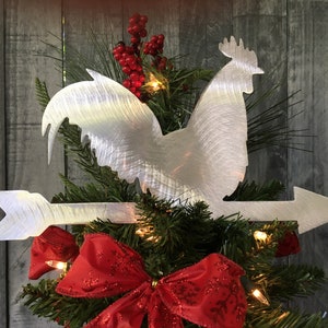 Puede incluir: Un gallo de metal plateado con una flecha apuntando hacia la derecha, en la cima de un &aacute;rbol de Navidad decorado con rojo y verde.
