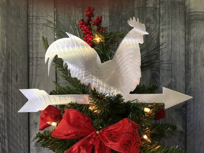 Puede incluir: Un gallo de metal plateado con una flecha apuntando hacia la derecha, en la cima de un &aacute;rbol de Navidad decorado con rojo y verde.
