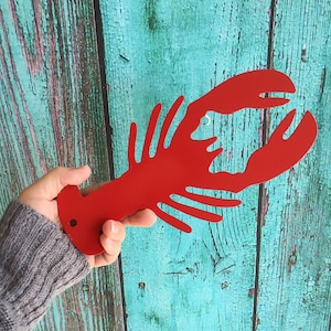 May include: A red metal lobster cutout, held in a hand against a teal blue wooden background.