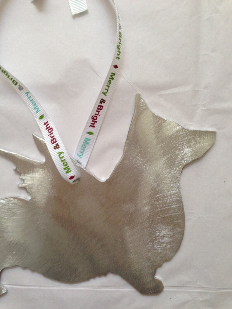 May include: Silver metal ornament shaped like a dog with a white ribbon attached. The ribbon has the words "Merry & Bright" printed on it.