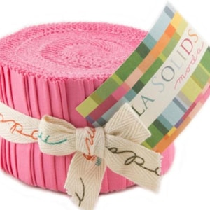 May include: A roll of pink fabric strips, bound with a cream-colored ribbon tied in a bow. The ribbon has decorative script. A colorful tag reads "LA SOLIDS moda". The fabric is tightly rolled and appears to be new.