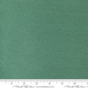 May include: A solid sage green felt fabric with a soft, matte, and slightly textured surface. A white ruler is visible at the bottom edge, showing measurements in inches from 1 to 7, with the text moda and www.modafabrics.com printed below.