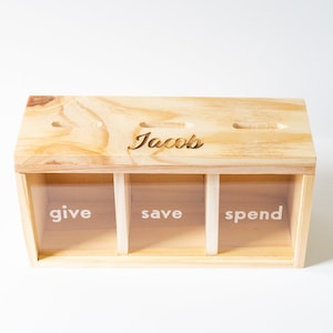 Puede incluir: Caja de madera con tres compartimentos etiquetados como "give", "save" y "spend". La caja tiene una ranura en la parte superior y est&aacute; personalizada con el nombre "Jacob's".
