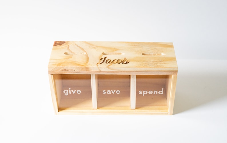 Puede incluir: Caja de madera con tres compartimentos etiquetados como "give", "save" y "spend". La caja tiene una ranura en la parte superior y est&aacute; personalizada con el nombre "Jacob's".