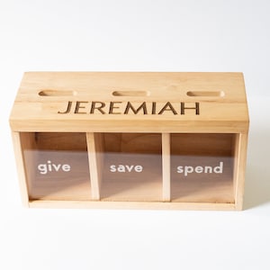 Puede incluir: Una caja de madera con tres compartimentos etiquetados como "give", "save" y "spend". La caja tiene una ranura en la parte superior y est&aacute; personalizada con el nombre "JEREMIAH".