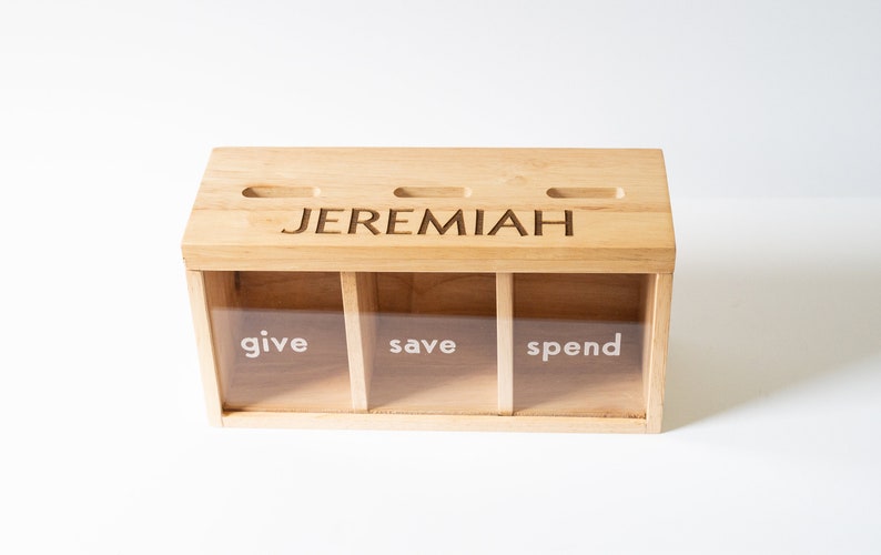 Puede incluir: Una caja de madera con tres compartimentos etiquetados como "give", "save" y "spend". La caja tiene una ranura en la parte superior y est&aacute; personalizada con el nombre "JEREMIAH".