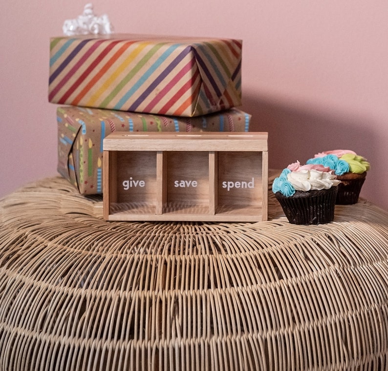 Puede incluir: Una caja de madera con tres compartimentos etiquetados como "give", "save" y "spend". La caja est&aacute; sobre una superficie tejida con tres cupcakes en el fondo.