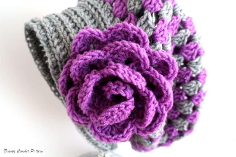 Pu&ograve; includere: Primo piano di un fiore all'uncinetto viola su uno sfondo all'uncinetto grigio. Il fiore &egrave; composto da pi&ugrave; strati di petali all'uncinetto.