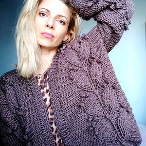 May include: A close-up of a woman wearing a purple crocheted cardigan with a cable knit pattern. The cardigan is open in the front and the woman is wearing a leopard print top underneath.