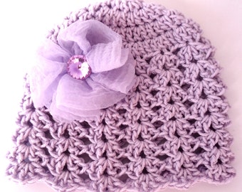 Puede incluir: Un gorro de crochet lavanda con una flor lavanda a juego y un brillante de cristal morado brillante en el centro.