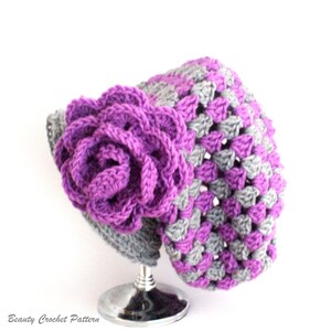 Pu&ograve; includere: Un cappello all'uncinetto viola e grigio con un grande fiore all'uncinetto viola sul lato. Il cappello &egrave; esposto su un supporto argentato.