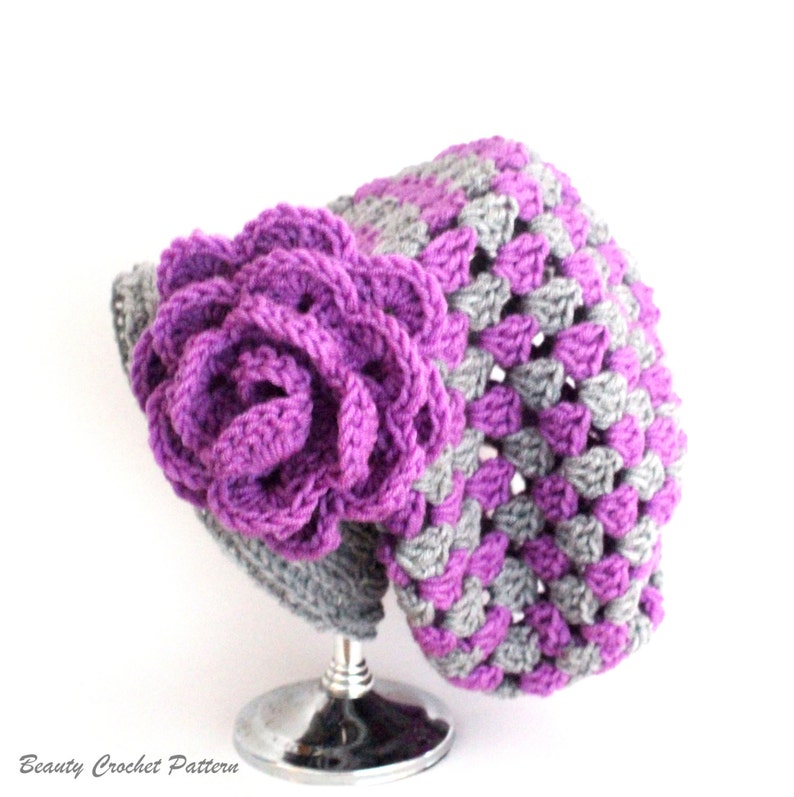 Pu&ograve; includere: Un cappello all'uncinetto viola e grigio con un grande fiore all'uncinetto viola sul lato. Il cappello &egrave; esposto su un supporto argentato.