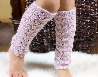 May include: A pair of light purple knitted leg warmers with a textured, openwork design. The leg warmers are made of a soft, possibly wool, material and are shown on a person's legs. The background includes a white rug and a wicker basket.