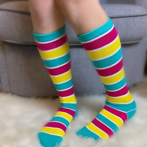Peut inclure: Des chaussettes montantes &agrave; rayures horizontales turquoise, roses, jaunes et blanches. Les chaussettes sont port&eacute;es sur des jambes, les pieds reposant sur une surface blanche et duveteuse. Les chaussettes ont un bord et un bout turquoise.