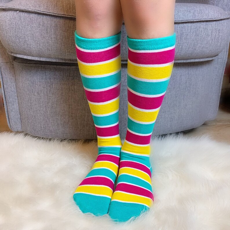 Peut inclure: Des chaussettes montantes &agrave; rayures horizontales turquoise, blanches, jaunes et magenta. Les chaussettes sont port&eacute;es par une personne et sont visibles des chevilles aux genoux. Le fond est un tapis doux et blanc.