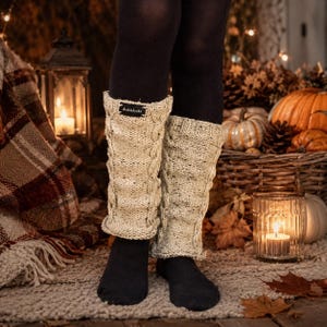 May include: A pair of beige cable-knit leg warmers featuring a speckled texture and a small black rectangular fabric tag with the white text Ankleknits sewn onto the upper edge of the left warmer, worn over solid black leggings and black socks.